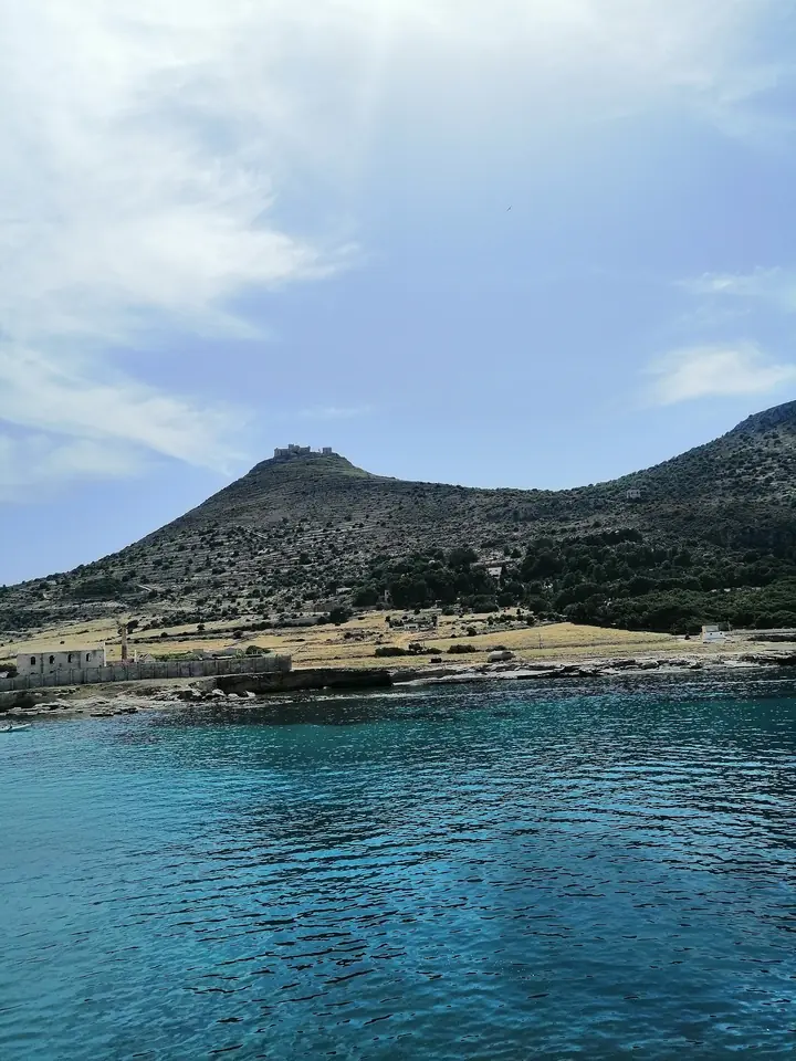 Blått hav vid Favignanas bebyggelse med berget och Castello di Santa Caterina ovanför