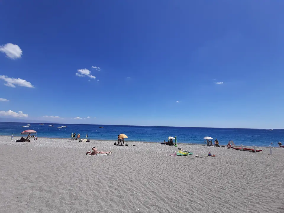 Grå strand, glest utspridda parasoller och blått hav i Mazzeo, som illustration till strandtyper